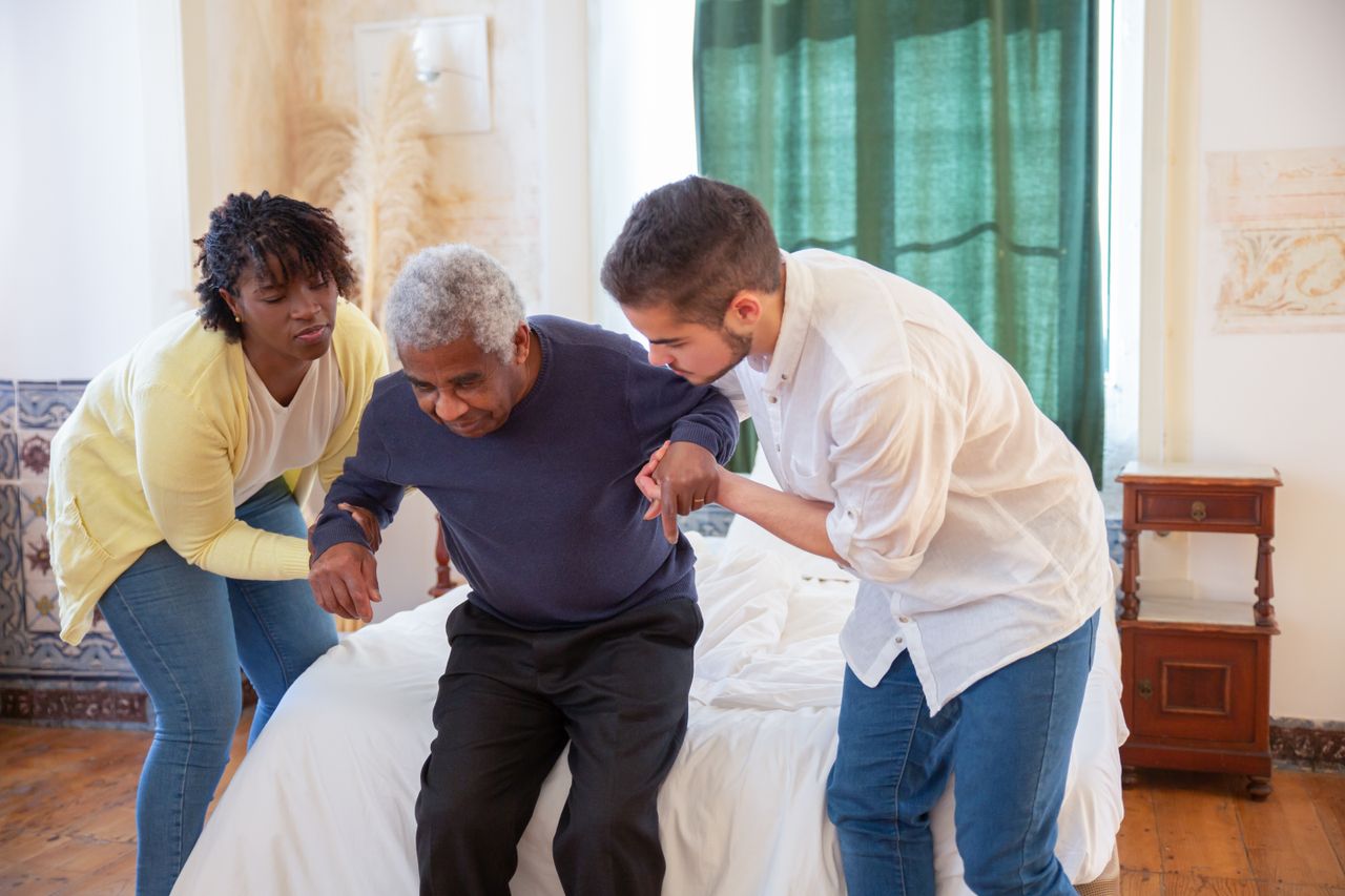 Home An elderly man receiving assistance from caregivers in a cozy home environment.